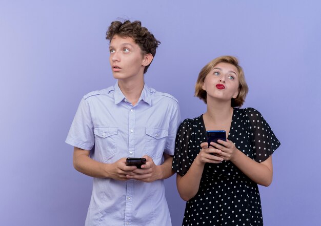 Jonge mooie paar man en vrouw met smartphones kijken verbaasd staande over blauwe muur