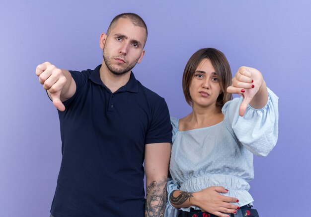 Jonge mooie paar man en vrouw ontevreden met duimen naar beneden staande over blauwe muur