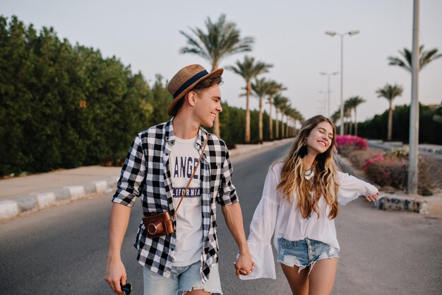 Jonge mooie paar op romantische outdoor date geniet van vrijheid en warme zomeravond in South City. Jongen in trendy geruit overhemd en meisje in vintage witte blouse lopen op de weg hand in hand