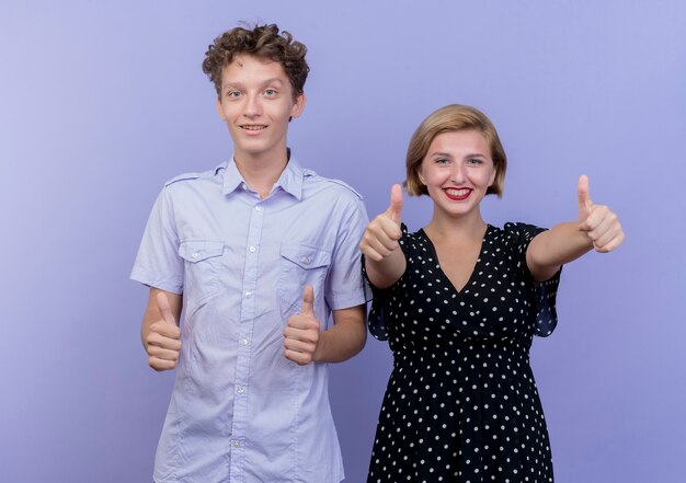 Jonge mooie paarman en vrouw die gelukkig en positief glimlachen tonen duimen die omhoog zich over blauwe muur bevinden