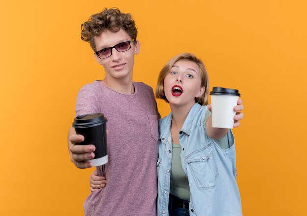 Jonge mooie paarman en vrouw die vrijetijdskleding dragen die koffiekopjes tonen die status over oranje muur glimlachen