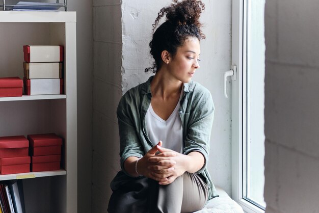 Jonge mooie peinzende vrouw met donker krullend haar zittend op de vensterbank in het publiek van de universiteit en bedachtzaam opzij kijkend