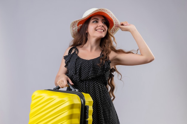 Jonge mooie reiziger meisje in jurk in polka dot in zomer hoed bedrijf koffer opzij glimlachend vrolijk blij en positief staande op witte achtergrond