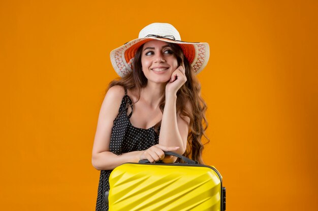 Jonge mooie reiziger meisje in jurk in polka dot in zomer hoed permanent met koffer glimlachend vrolijk met blij gezicht op gele achtergrond