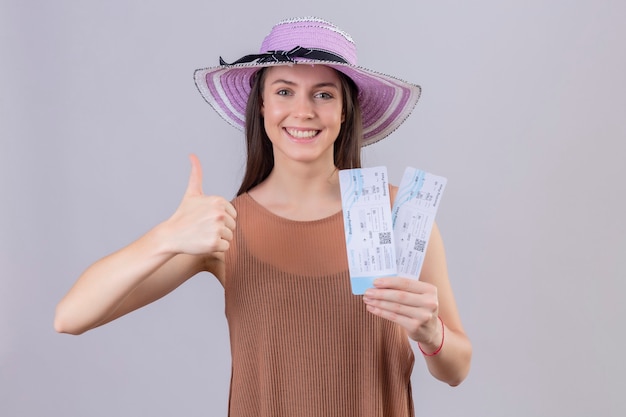 Jonge mooie reiziger vrouw in zomer hoed bedrijf vliegtickets lachend met blij gezicht duimen opdagen staande op witte achtergrond