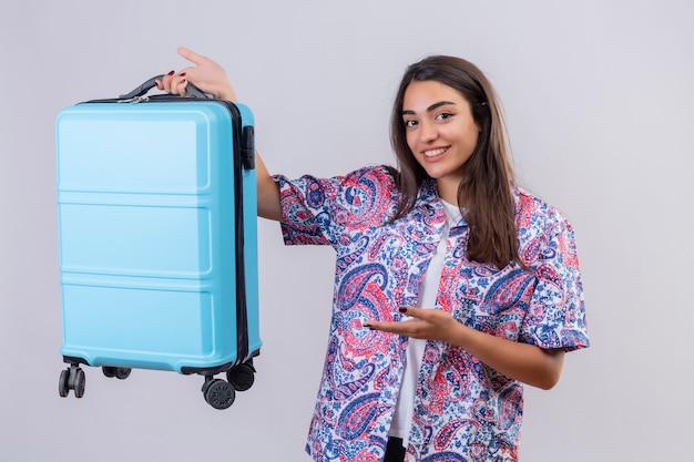 Jonge mooie reiziger vrouw met blauwe koffer wijzend met arm van de hand naar het glimlachend vrolijk staande op witte achtergrond