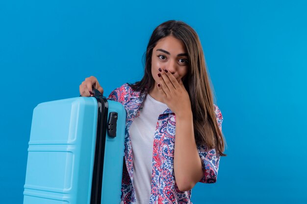 Jonge mooie reizigersvrouw die blauwe koffer houdt die verbaasd en verbaasd kijkt die mond behandelt met hand die zich over blauwe achtergrond bevindt