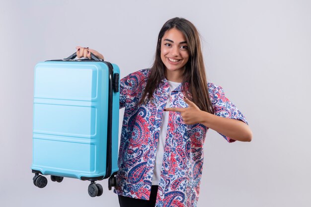 Jonge mooie reizigersvrouw die blauwe koffer houdt richtend met wijsvinger aan het vrolijk glimlachend over witte muur