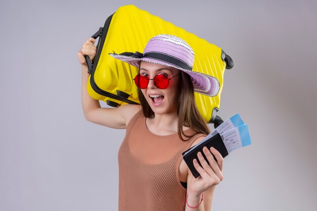 Jonge mooie reizigersvrouw die in de zomerhoed rode zonnebril dragen die gele koffer en luchtkaartjes houden die vrolijk met gelukkig gezicht over witte muur glimlachen