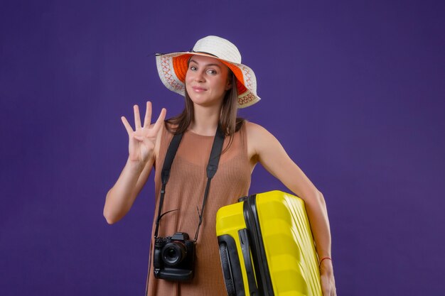 Jonge mooie reizigersvrouw in de zomerhoed met gele koffer en camera positief en gelukkig glimlachend tonend nummer drie over purpere muur