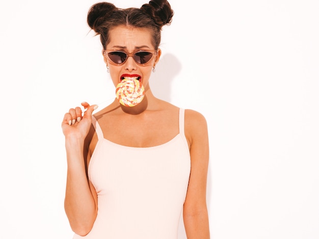 Gratis foto jonge mooie sexy hipster vrouw met rode lippen in zonnebril. trendy meisje in zomer badmode kleding. eten, bijten snoep lolly