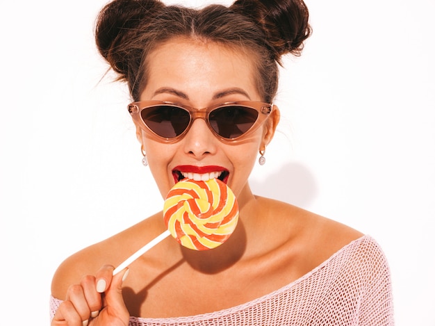 Gratis foto jonge mooie sexy hipster vrouw met rode lippen in zonnebril. trendy meisje in zomer badmode kleding. snoeplolly eten