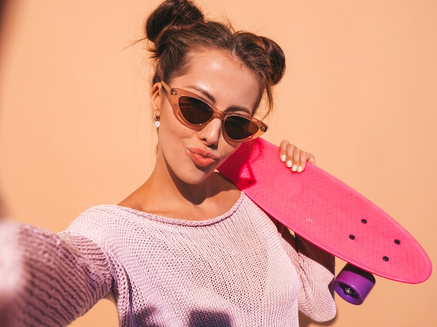 Jonge mooie sexy lachende hipster vrouw in zonnebril. trendy meisje in zomer gebreid vest. vrouw met roze penny skateboard, geïsoleerd op beige muur. selfie zelfportret foto's maken op phon