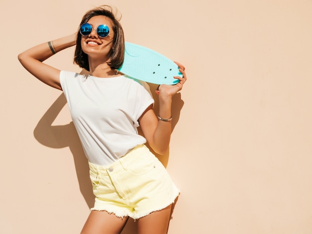Jonge mooie sexy lachende hipster vrouw in zonnebril. Trendy meisje in zomer T-shirt en korte broek. Positieve vrouw met blauwe cent skateboard poseren in de straat in de buurt van muur