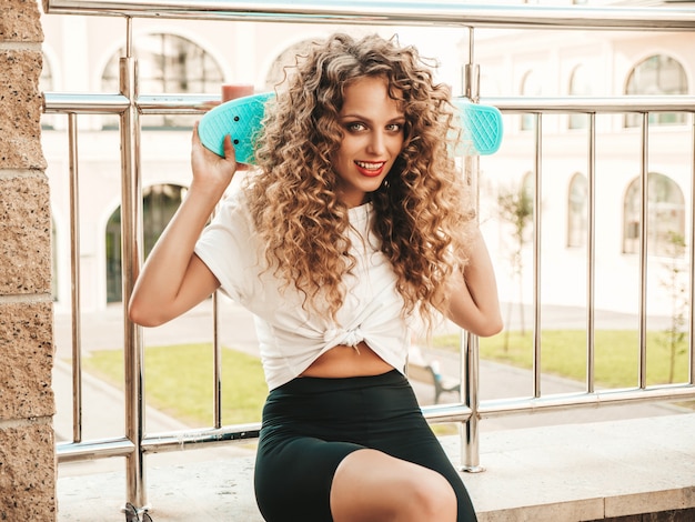 Jonge mooie sexy lachende hipster vrouw met afro krullen kapsel. Trendy meisje in zomerkleren. Positief wijfje met blauwe stuiver skateboard zittend op de straat achtergrond