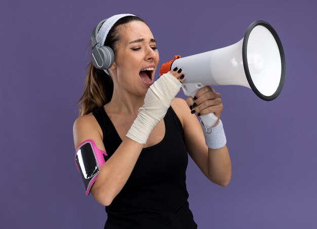 Jonge mooie sportieve meisje dragen hoofdband polsbandjes koptelefoon en telefoon armband met gewonde pols gewikkeld met verband praten door spreker geïsoleerd op paarse muur
