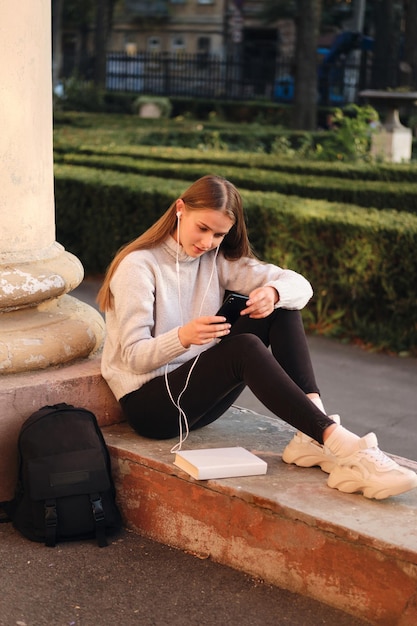Jonge mooie stijlvolle student meisje in gezellige trui dromerig met behulp van mobiele telefoon tijdens studiepauze buiten
