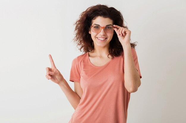 Jonge mooie stijlvolle vrouw in glazen met vinger, krullend haar, glimlachen, positieve stemming, gebaar, blij emotie, geïsoleerd, roze t-shirt