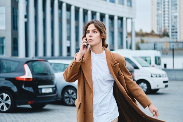 Jonge mooie stijlvolle vrouw in jas met mobiele telefoon die vol vertrouwen door de straat van de stad loopt