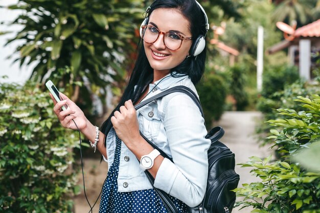 Jonge mooie stijlvolle vrouw met behulp van smartphone, hoofdtelefoon, bril, zomer, vintage denim outfit, glimlachen, gelukkig, positief