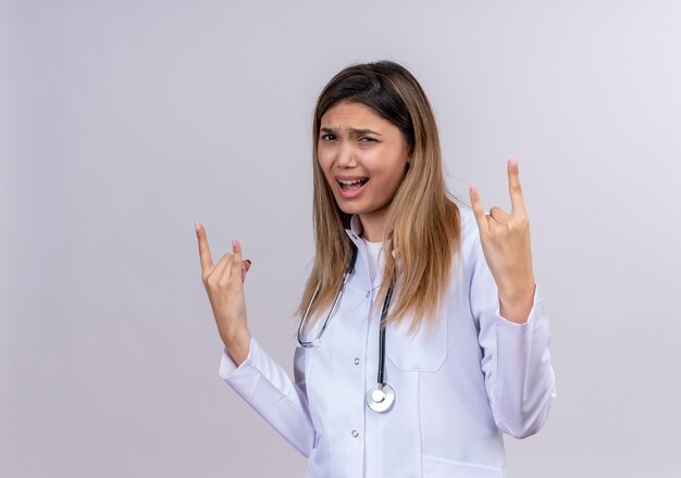 Jonge mooie vrouw arts die witte jas met een stethoscoop draagt die zelfverzekerd kijkt en rotssymbolen doet met vingers van beide handen