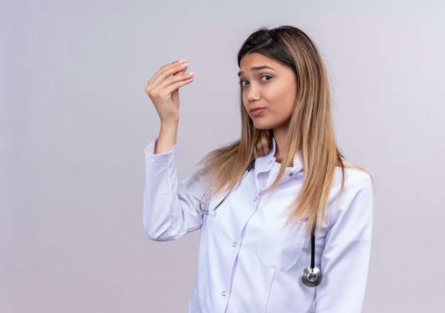 Jonge mooie vrouw arts die witte jas met stethoscoop draagt, die vinger wrijft die geldgebaar met hand maakt die om geld vraagt