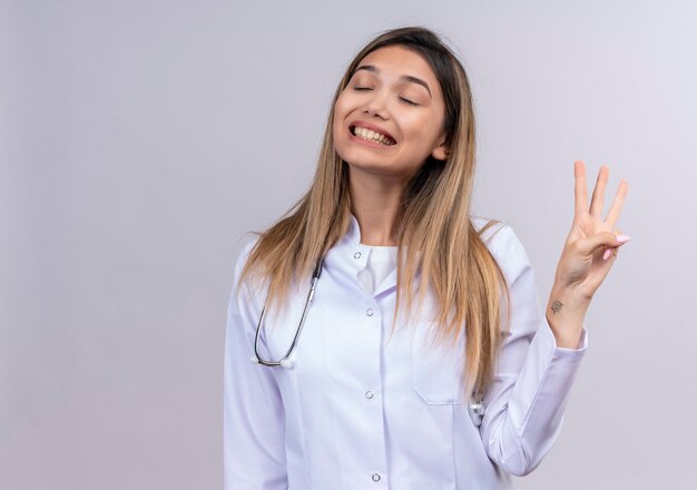 Jonge mooie vrouw arts die witte jas met stethoscoop met gesloten ogen draagt die vrolijk nummer drie met vingers toont