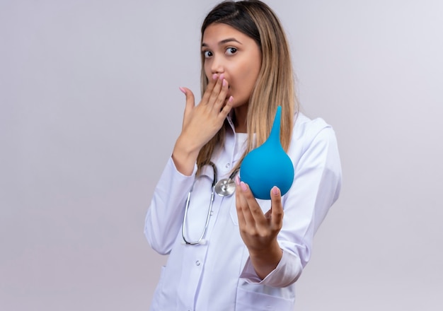 Jonge mooie vrouw arts die witte laag met stethoscoop draagt die een klysma houdt die geschokt kijkt die mond behandelt met hand