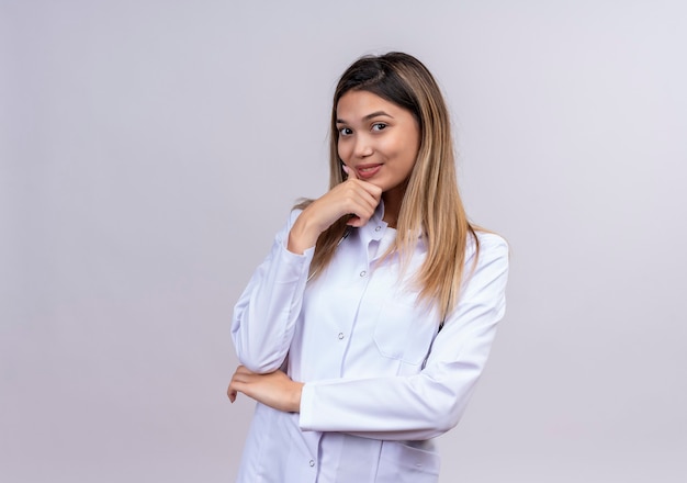 Jonge mooie vrouw arts die witte laag met stethoscoop draagt die glimlachend positief en gelukkig status met hand op kin kijkt