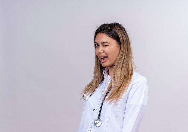 Jonge mooie vrouw arts dragen witte jas met een stethoscoop knipogen en glimlachen