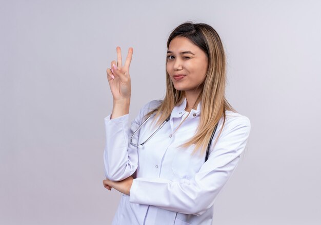 Jonge mooie vrouw arts dragen witte jas met stethoscoop glimlachend en knipogen weergegeven: overwinningsteken