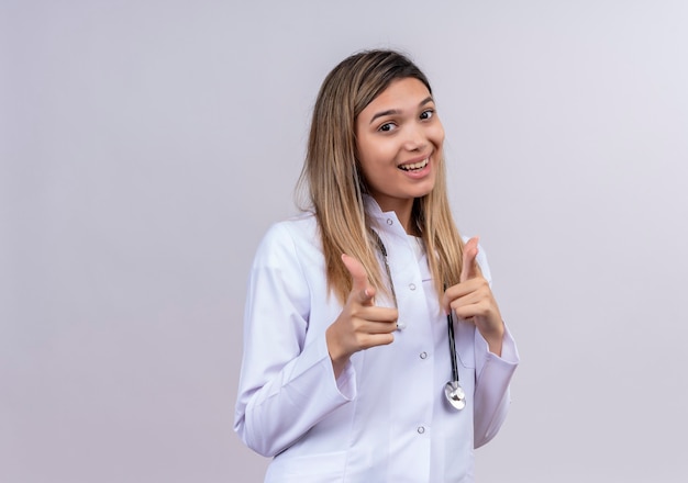 Jonge mooie vrouw arts dragen witte jas met stethoscoop glimlachend vrolijk wijzende wijsvingers