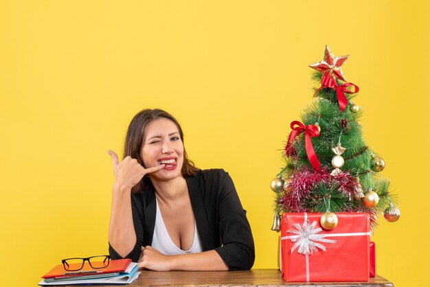 Jonge mooie vrouw bellen me gebaar zittend aan een tafel in de buurt van versierde kerstboom op kantoor op geel