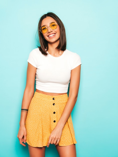 Gratis foto jonge mooie vrouw die camera bekijkt. trendy meisje in casual zomer wit t-shirt en gele rok in ronde zonnebril. positieve vrouw toont gezichtsemoties. grappig model dat op blauw wordt geïsoleerd