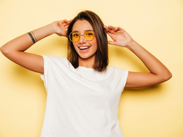 Jonge mooie vrouw die camera bekijkt. trendy meisje in casual zomer wit t-shirt en jeans short in ronde zonnebril. positieve vrouw toont gezichtsemoties. grappig model dat op geel wordt geïsoleerd