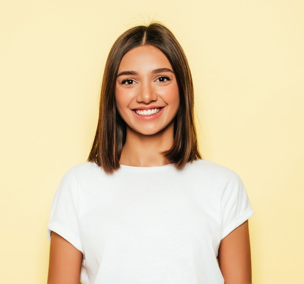 Jonge mooie vrouw die camera bekijkt. Trendy meisje in casual zomer wit T-shirt en jeans short. Positieve vrouw toont gezichtsemoties. Grappig model dat op geel wordt geïsoleerd