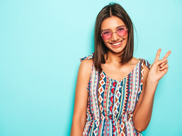 Jonge mooie vrouw die camera bekijkt. Trendy meisje in casual zomerjurk en ronde zonnebril. Positieve vrouw toont gezichtsemoties. Grappig model dat op blauw wordt geïsoleerd