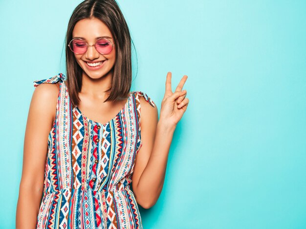 Jonge mooie vrouw die camera bekijkt. Trendy meisje in casual zomerjurk en ronde zonnebril. Positieve vrouw toont gezichtsemoties. Grappig model dat op blauw wordt geïsoleerd