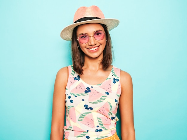 Jonge mooie vrouw die camera in hoed bekijkt. trendy meisje in casual zomer wit t-shirt en gele rok in ronde zonnebril. positieve vrouw toont gezichtsemoties. grappig model dat op blauw wordt geïsoleerd