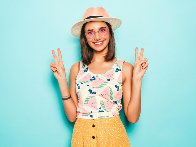 Jonge mooie vrouw die camera in hoed bekijkt. Trendy meisje in casual zomer wit T-shirt en gele rok in ronde zonnebril. Positieve vrouw toont gezichtsemoties. Vredesteken toont