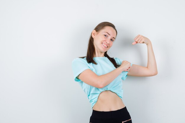 Jonge mooie vrouw die de spieren van de arm toont, met de hand op de arm in een t-shirt en er trots uitziet, vooraanzicht.