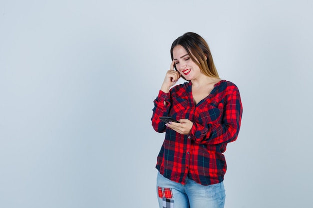 Jonge, mooie vrouw die de telefoon vasthoudt, de hand op het gezicht houdt in een casual shirt en er vrolijk uitziet, vooraanzicht.