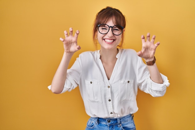Jonge mooie vrouw die een casual shirt draagt over een gele achtergrond die grappig klauwgebaar doet als agressieve en sexy uitdrukking van een kat