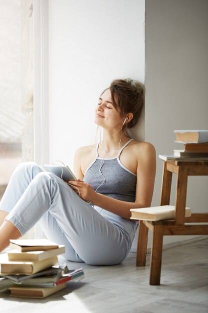 Jonge mooie vrouw die in hoofdtelefoons met gesloten ogen glimlachen die tablet houden luisterend aan stromende muziekzitting op vloer over witte muur.