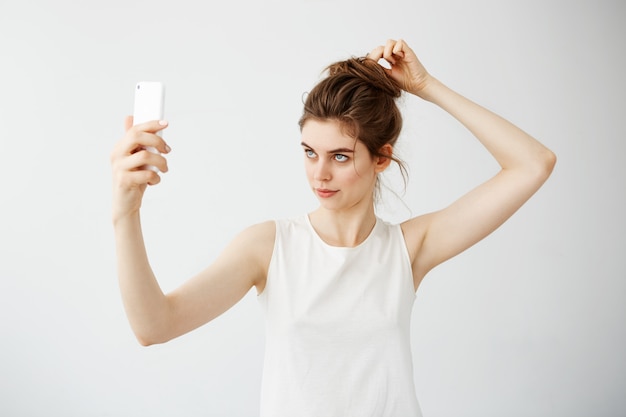 Jonge mooie vrouw die met broodje het corrigerende haar van het telefoonscherm over witte achtergrond bekijkt.