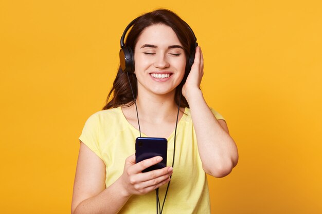 Jonge mooie vrouw die met hoofdtelefoons aan favoriete muziek luistert die op geel wordt geïsoleerd