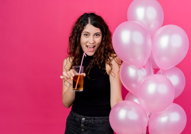 Jonge mooie vrouw die met krullend haar bos van luchtballons en cocktail houdt die vrolijk over roze muur glimlacht