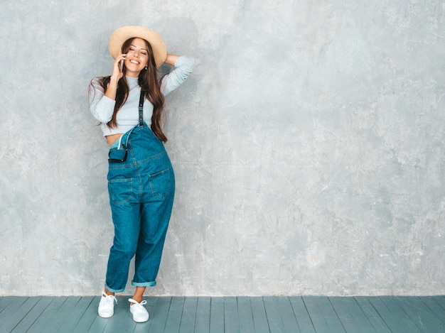 Jonge mooie vrouw die op telefoon spreekt. Trendy meisje in casual zomer overall kleding en hoed.