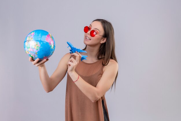 Jonge mooie vrouw die rode zonnebril dragen die bol en stuk speelgoed vliegtuig speels en gelukkig over witte muur houden