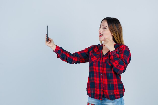 Jonge mooie vrouw die selfie neemt, met de hand in de buurt van het gezicht in een casual shirt en er vrolijk uitziet, vooraanzicht.